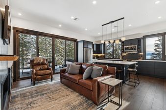 A modern open-concept living and kitchen area in a mountain home features a brown leather sofa, an armchair, a kitchen island with bar stools, and large windows revealing a snowy forest.