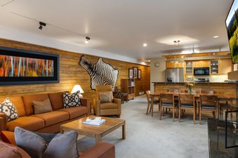 An expansive open-concept living and dining area in Montaneros 202, featuring an orange sofa, a wooden coffee table, a zebra hide on the wall, and a modern kitchen.
