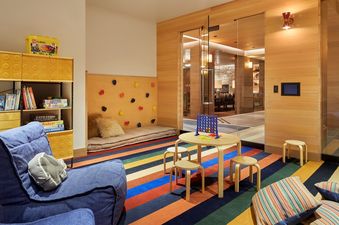 A vibrant games room in the Caldera House Four Bedroom Suite features a colorful striped rug, a wooden table with Connect Four, a padded climbing wall, and a Lego-inspired storage unit with games and books.