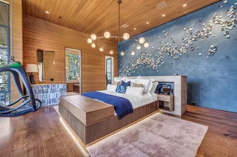A luxurious and modern master bedroom in Monitors Rest featuring a large bed with a blue blanket, a dark blue accent wall with sculptural art, and wooden paneling throughout.