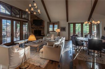 A spacious great room at Martin Creek Cottage features a cozy living area with a stone fireplace and a dining area with a large table, all illuminated by elegant chandeliers and natural light.