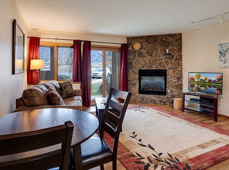 A wide shot of the living and dining area in The Pines Condominiums P103C, featuring a brown sofa, a wooden dining table, a stone fireplace, and a television.