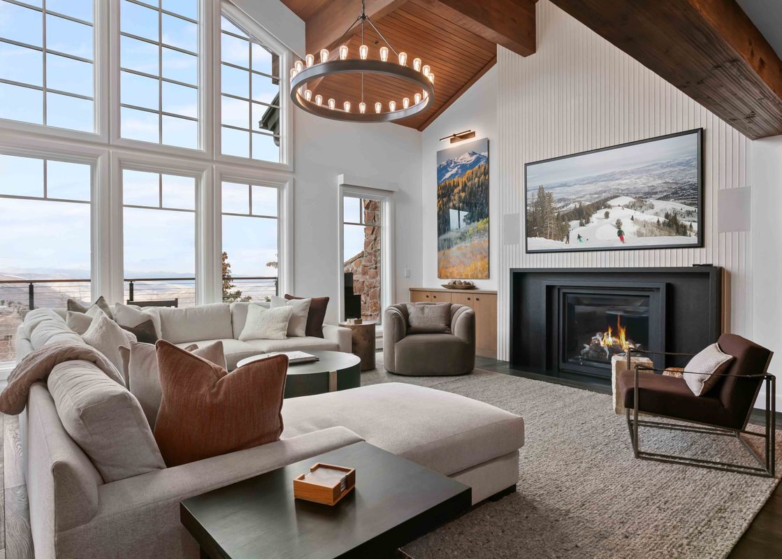 A spacious and modern living room at Stag Lodge 11 4BR with a large sectional sofa, a black fireplace, a widescreen TV, mountain view artwork, and tall windows overlooking a scenic mountain landscape.