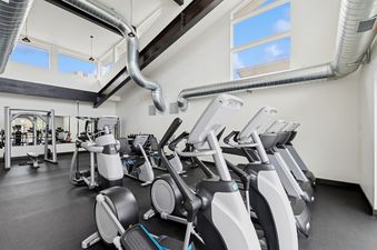 A modern and spacious fitness center at Quarry A28 with a variety of exercise equipment, high ceilings, large windows, and exposed ductwork.