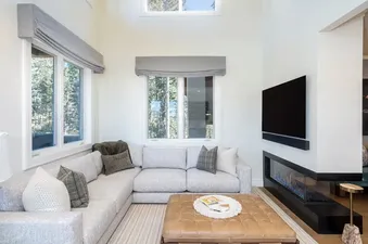 A modern living room with a large sectional sofa, a leather ottoman, a wall-mounted TV, and a linear fireplace with a view of trees outside the windows.