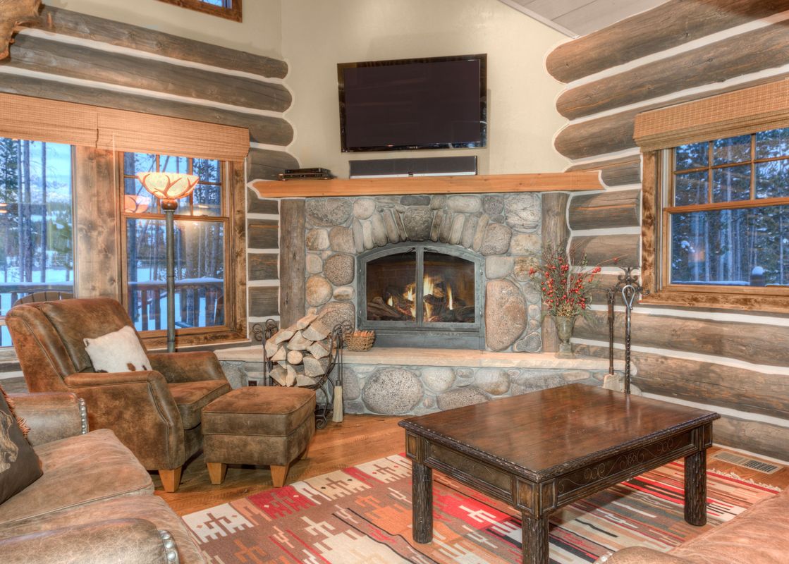 A cozy, rustic living room with a large stone fireplace, comfortable leather furniture, and a view of a snowy forest through the windows.