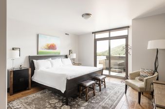 A luxurious king bedroom with a dark wood bed, white bedding, and two leather stools, featuring a vibrant tree painting, modern nightstands, elegant lamps, an upholstered armchair, and a glass door opening to a balcony with mountain views and a hot tub.