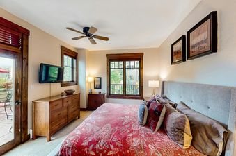 A king bedroom in Water House On Main Street 5205 with a large bed, a ceiling fan, a wall-mounted TV, and a view of the outdoors through several windows.