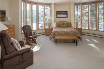 A cozy bedroom at Hillside Hideaway featuring a large bed, comfortable seating, and panoramic windows with views of snow-covered mountains and pine forests.