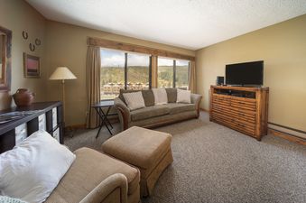 A cozy living room in Snowblaze E310 with a comfortable sofa, armchair, ottoman, and a large window offering scenic views of the mountains and resort.