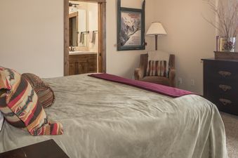 A cozy bedroom at 60 Moose Ridge features a large bed with a colorful throw, an armchair, and an en-suite bathroom visible through an open doorway.