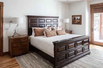 The bedroom in Moonlight Basin Luxury Suite 6A has a king-sized bed with a wooden headboard and a white duvet, white, tan, and orange pillows, two nightstands with lamps, a rug, and a door to the outside.