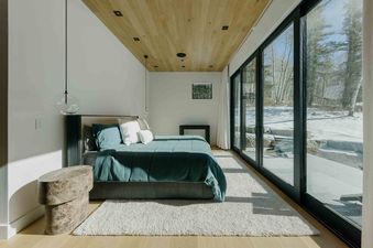 A bedroom with a king-sized bed, a white rug, and a large window with a view of a snowy forest.