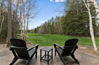 A peaceful outdoor patio features two black Adirondack chairs and a matching side table, overlooking a vibrant green golf course and a lush forest under a clear blue sky.