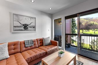A cozy living room in Snowcrest 216 with a brown leather sofa, longhorn bull artwork, and a balcony with mountain views.