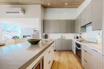 A modern kitchen with light gray cabinets, white countertops, stainless steel appliances, and a large central island with a green bowl. The kitchen seamlessly connects to an open living area with large windows.