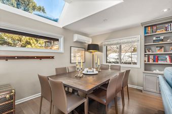 The dining room at Dolomite Villas Unit 10 features a square wooden table with eight brown leather chairs, illuminated by natural light from two large windows and a ceiling skylight.
