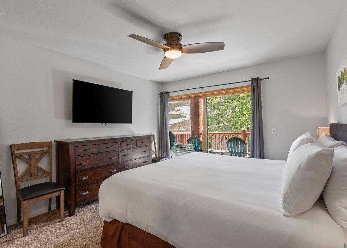 A master bedroom with a king-size bed, a large flat-screen TV above a wooden dresser, a ceiling fan, and a private balcony overlooking a scenic landscape with trees and snow.