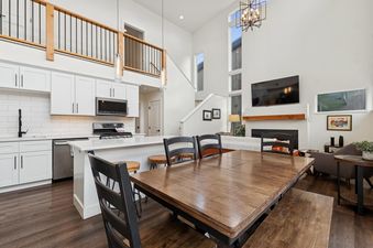 A modern open-concept kitchen and living area with white cabinetry, a large dining table, and a living room with a fireplace and high ceilings.