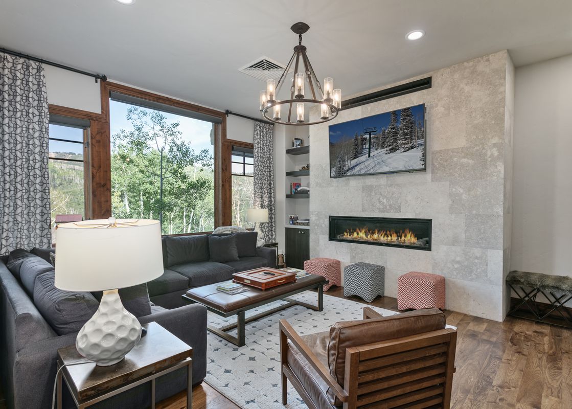 A modern and luxurious living room at Silver Strike Lodge 407 with a gray sectional sofa, a leather armchair, a contemporary coffee table, a geometric rug, a chandelier, and a flat-screen TV above a fireplace, all set against a backdrop of large windows w