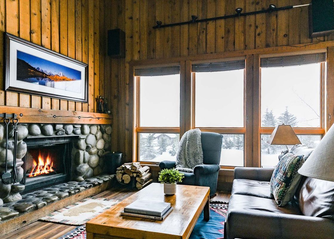 Cozy living room in Nez Perce A6 with a stone fireplace, leather sofa, and snow-covered trees visible through large windows.