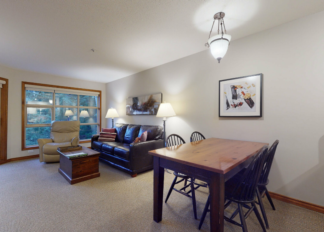 The living area within Aspens on Blackcomb Unit 415 features a comfortable dark leather sofa and a recliner, alongside a wooden coffee table and a dining table with chairs, all set on a light-colored carpet.