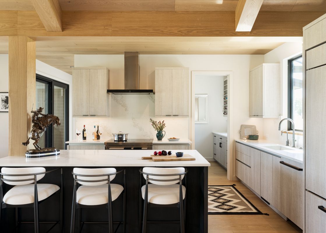 A modern kitchen with light wood cabinets, white countertops, and a large island with three bar stools, featuring a cowboy sculpture.