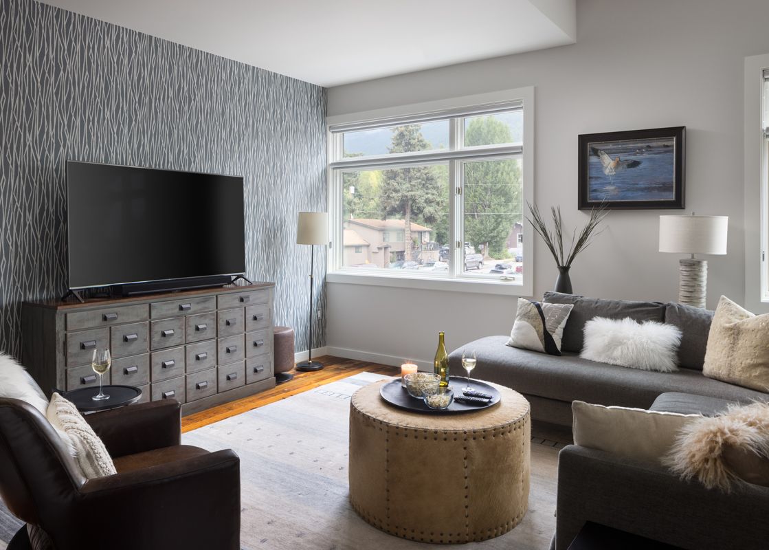 A modern and inviting living room in Pied a Terre 202 featuring a large TV, an L-shaped sofa, a round ottoman, and natural light from a window with an exterior view.