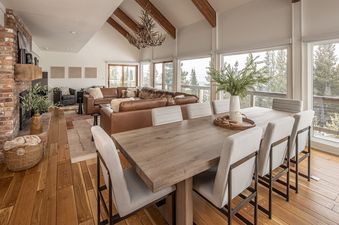 A spacious great room with a dining area and living room, featuring a large fireplace, vaulted ceilings, and panoramic windows overlooking a snowy forest.