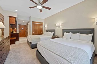 A hotel bedroom featuring two large beds with white bedding, a wooden dresser, and a ceiling fan, illuminated by wall sconces and recessed lighting.