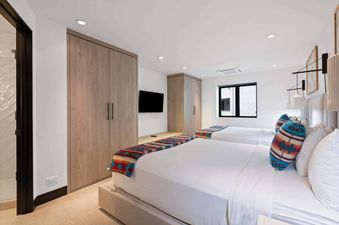 A modern bedroom with two single beds, each with white linens and a colorful throw, two wooden wardrobes, a wall-mounted TV, and a large window.