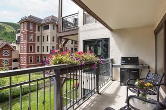 The private balcony at Montaneros 314 features colorful flower boxes, a BBQ grill, and comfortable seating, all set against a backdrop of scenic mountain architecture and lush greenery.
