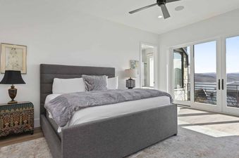 A spacious bedroom at SkyRidge 1245 showcasing a large gray bed with plush bedding, two distinctive nightstands with lamps, and expansive glass doors opening to a balcony with scenic mountain and water views.
