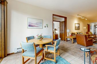 The dining area of SunRiver E101 features a round table with four chairs, a large artwork on the wall, and a view into the living room.