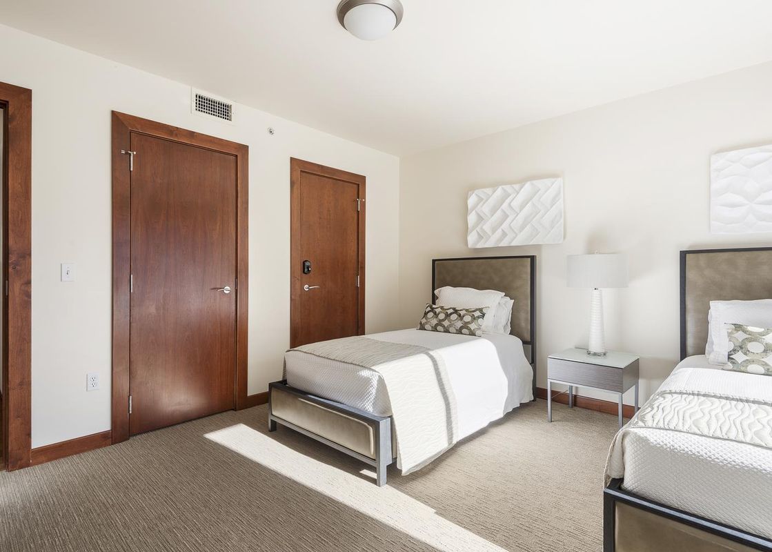 Two twin beds with white linens and patterned pillows in a light and airy bedroom with wood floors and large windows.