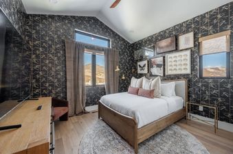 A modern bedroom with dark patterned wallpaper, a wooden bed, and windows looking out onto the mountains.