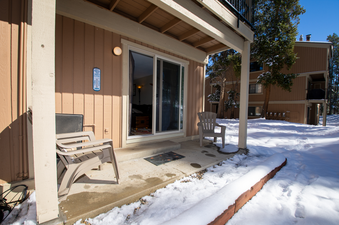 The ground-level patio of Beaver Village 211 is shown covered in snow, featuring two plastic chairs, a sliding glass door, and an exterior light fixture on the building.