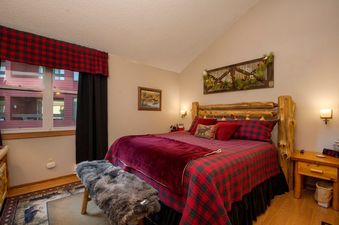A cozy bedroom with a large bed, featuring a rustic wooden headboard and red plaid bedding, is bathed in warm light.