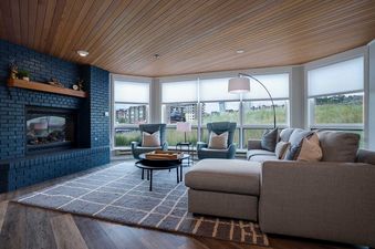 A modern living room with a dark blue brick fireplace, grey sectional sofa, and two blue armchairs, featuring panoramic windows with views.