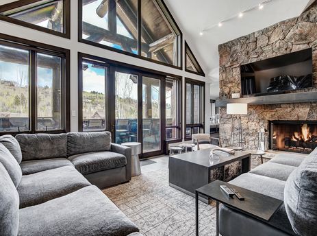 A spacious living room at Snow Cloud Lodge 506 features a large grey sectional sofa, a dark coffee table, and a stone fireplace with a mounted TV, all set against a backdrop of large windows with scenic outdoor views.