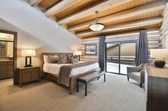 A rustic bedroom featuring a large wooden bed, log cabin walls, and a beamed ceiling, with a glass door opening to a snowy outdoor view.