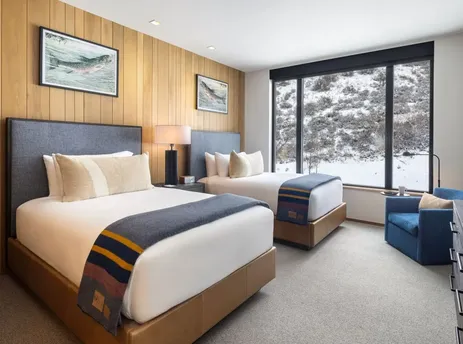 Two beds with white bedding, patterned throws, and blue headboards in a room with a wood-paneled wall, large window with a snowy view, and a blue armchair.