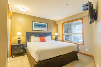 A spacious bedroom at Aspens on Blackcomb Unit 458 features a king-sized bed with white linens and a red accent pillow, flanked by two dark nightstands with matching lamps. A large window with blinds offers an exterior view, and a wall-mounted TV is visib