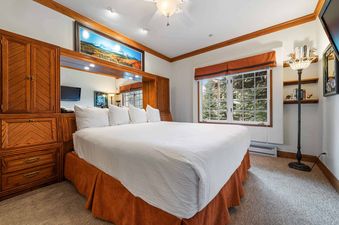 A cozy bedroom at The Charter E0405 with a king-sized bed, wooden furnishings, a window view of pine trees, and warm lighting.