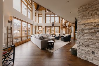 A wide shot of High West at the Plaza's living room showcasing its spacious layout, numerous windows with mountain views, a large gray sofa, two black leather armchairs, a stone fireplace, and a wooden spiral staircase.