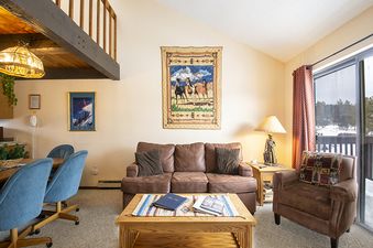 The living area features a brown sofa and armchair, a wooden coffee table, a dining table with blue chairs, and a large window looking out onto a snowy landscape.