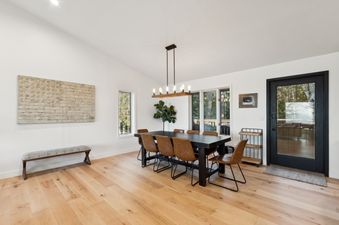 A modern dining room featuring a dark wooden table with eight brown leather chairs, light wood floors, and large windows with outdoor views.
