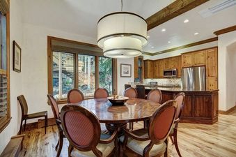 A large, bright dining area featuring a round wooden dining table surrounded by cushioned chairs, with a view of a modern kitchen and an exterior through large windows.