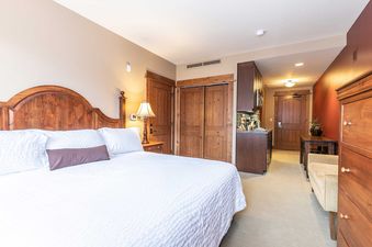 A hotel room with a king size bed, a brown dresser and a closet, a small kitchenette and a hallway with a door to the outside.