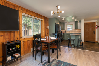The dining area and kitchen of Beaver Village 514 are visible, featuring a wooden dining table set for four, surrounded by black chairs, next to a large window offering an exterior view of trees.